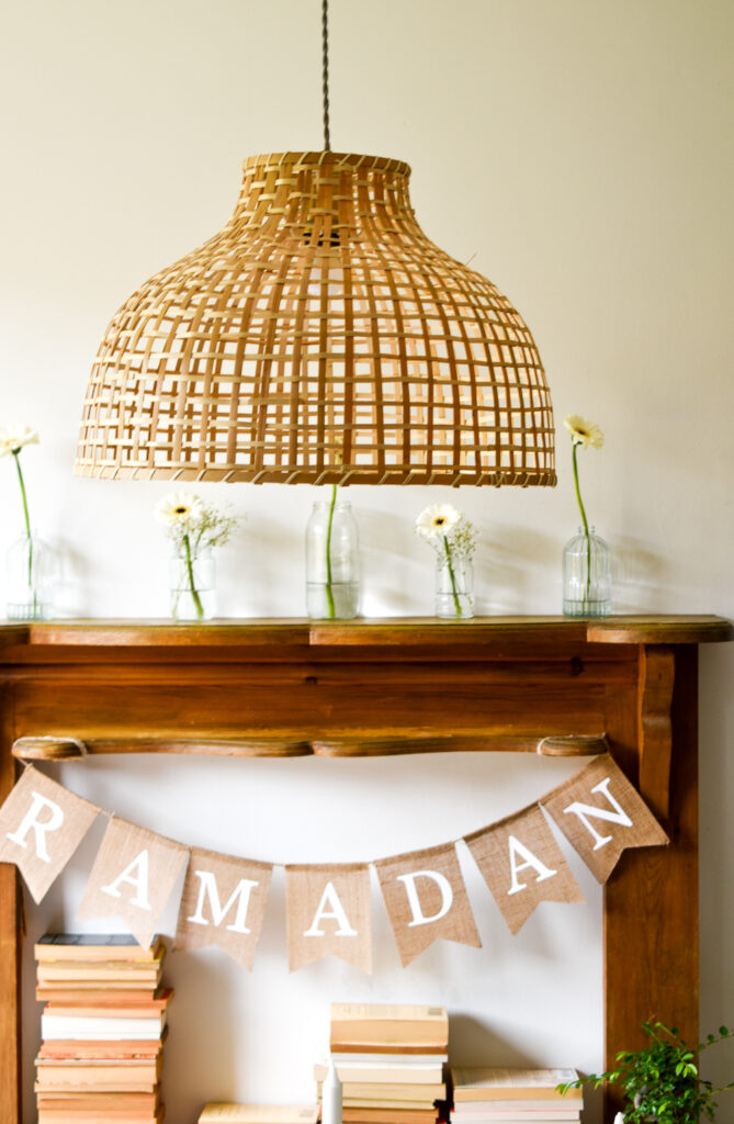 a large wicker light shade is centre top of the photo. Gerberas which are white/cream with a black centre stand at different heights, spaced out in glass jars. They sit on a faux fireplace mantle which is wooden. A hessian bunting with white printed letters reads Ramadan and piles of books sit bottom of the photo. A small green plant is bottom right. 