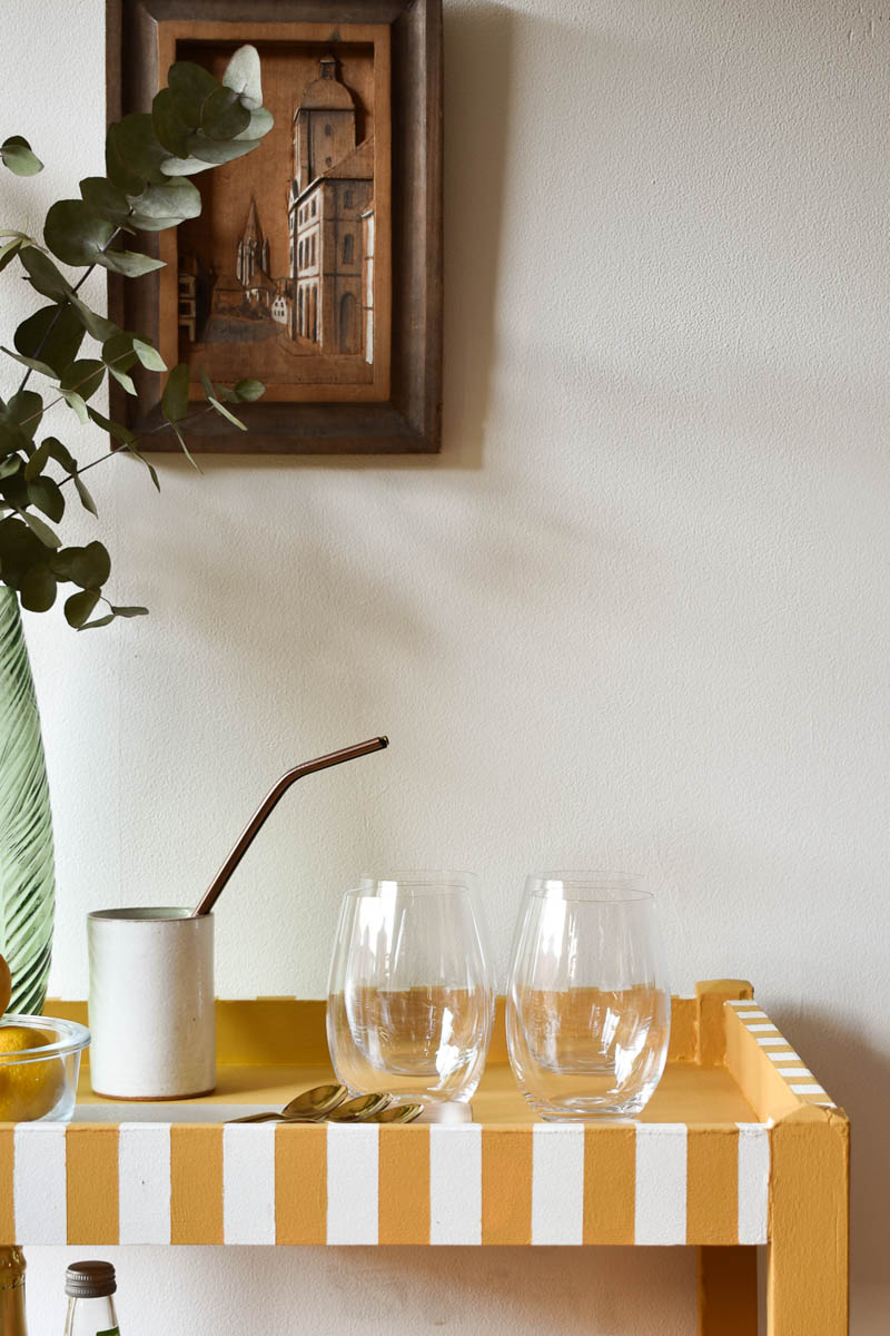 DIY bar cart with glasses and white decanter with a straw next to vase with greenery