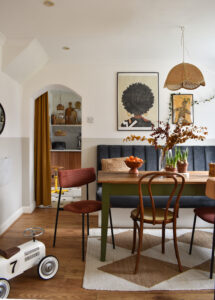 a vintage modern dining room with warm tones. A painting of a Black woman is hung on the wall above the banquet seating. Her afro is perfectly rounded. A rattan vintage lampshade hangs above a vintage table with a sanded top and dark green legs.