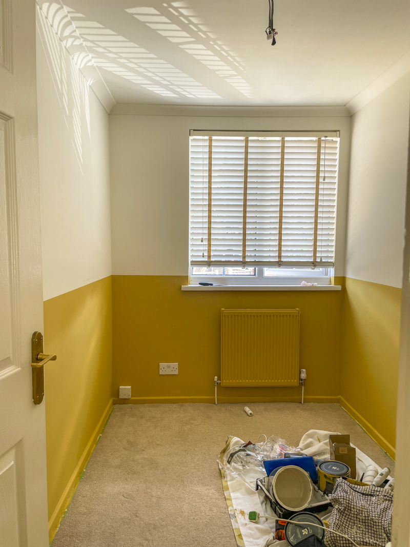 a half painted room finished, a pile of painting equipment lays bottom right on a dust sheet on a beige carpet. 