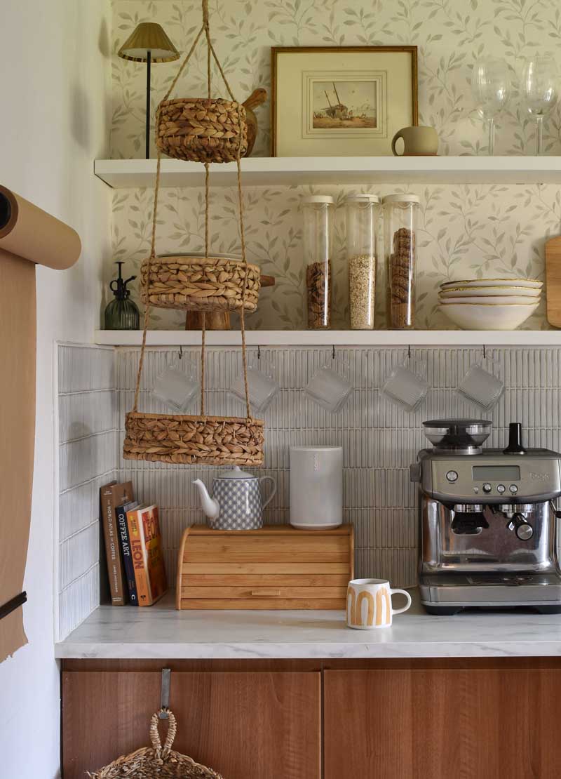 Open kitchen shelving styled with plates, glassware and jars of cereal. Beneath is a faux marble worktop with a coffee maker, a bread bin, books and a teapot. Glass mugs are hanging from the lower shelf, and there is a three-tiered storage basket hanging from the ceiling in front. The wall behind the shelves is papered in a soft leaf print, and a wall-mounted roll of brown paper is just visible to one side.