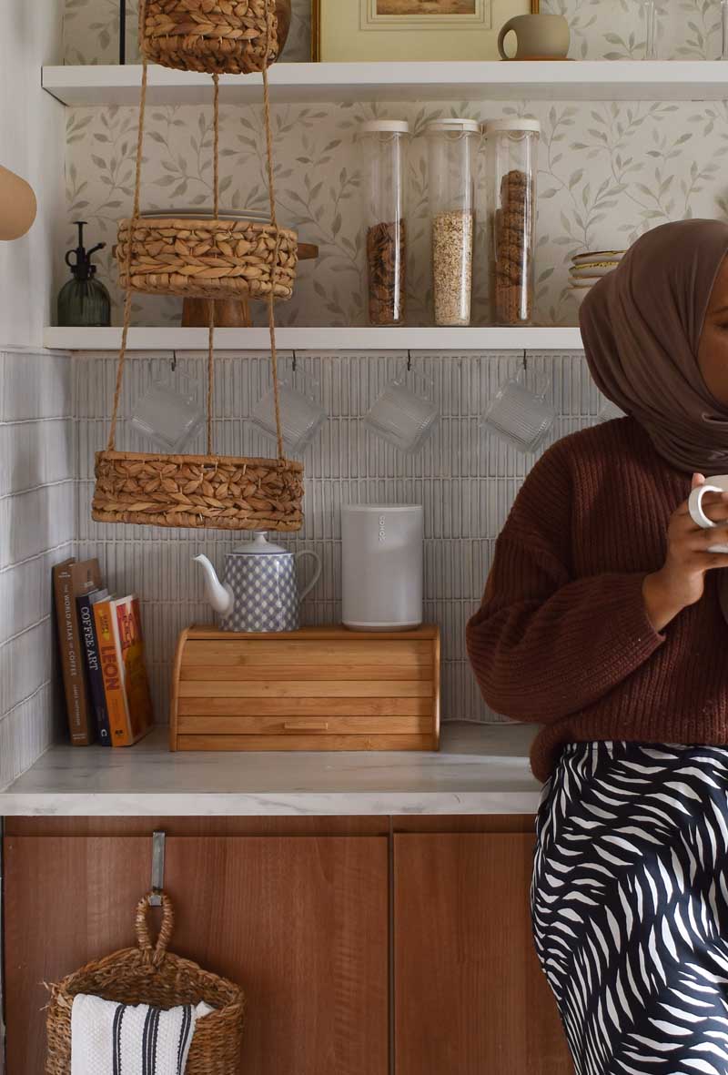 Open kitchen shelving styled with plates, glassware and jars of cereal. Beneath is a faux marble worktop with a bread bin, books and a teapot. Medina is partially visible, standing to one side and holding a cup of coffee.