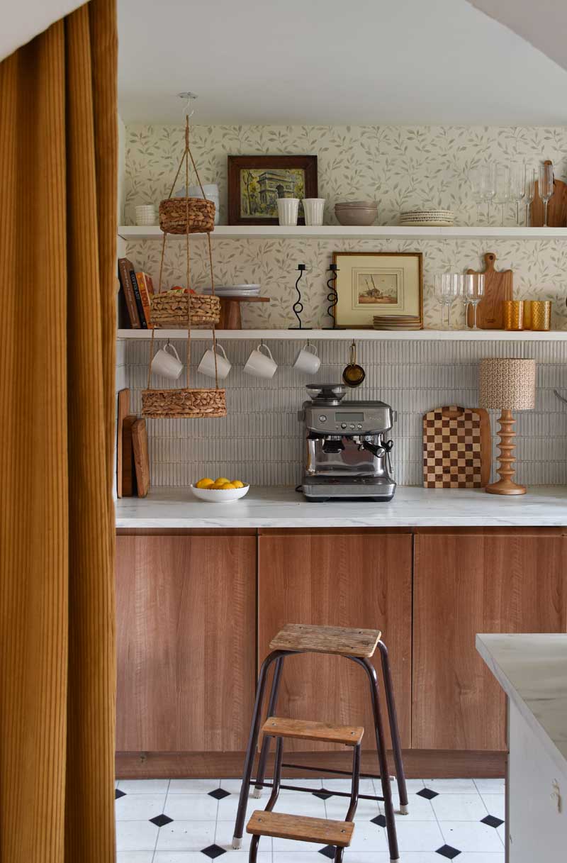 A peek into the kitchen through a mustard yellow thick ribbed curtain. Open shelving with plates, glassware, a cake stand and cutting boards sits above a faux marble worktop. There is a coffee maker, a bowl of fruit and a wavy-based lamp on the worktop, and mugs hanging from the lower shelf.