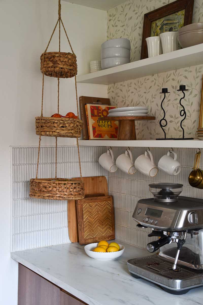 A pair of open kitchen shelves styled with plates, a wooden cake stand, books, glassware and wavy candlesticks. Beneath is a faux marble worktop with a coffee maker, chopping boards and a bowl of fruit. Mugs are hanging from the lower shelf, and there is a three-tiered storage basket hanging from the ceiling in front of them.