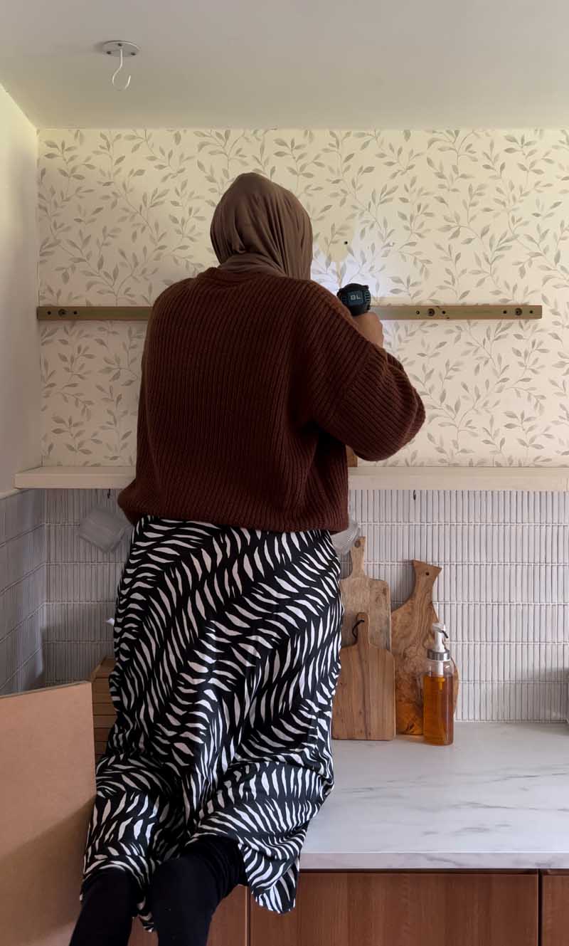 Medina is installing open shelving above wooden kitchen units, a faux marble worktop and a tiled splashback. Patterned wallpaper covers the wall behind.