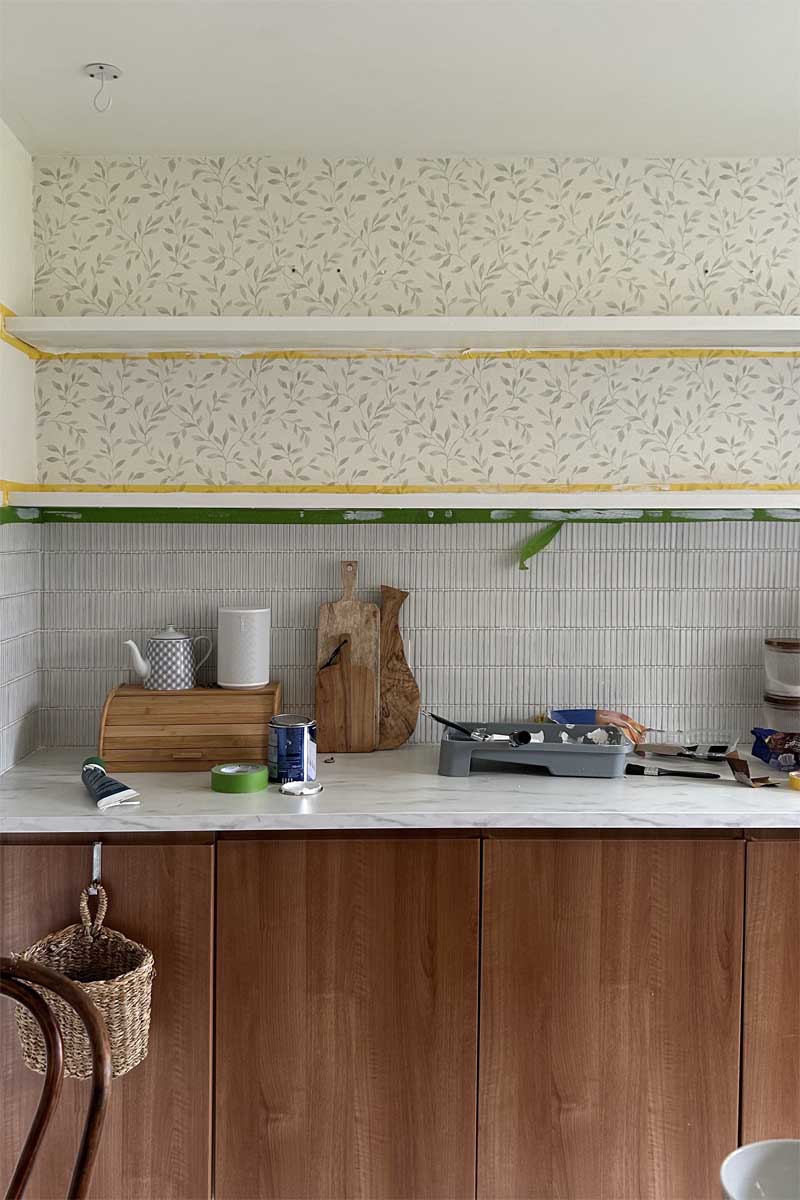 Newly installed open shelving above wooden kitchen units, a faux marble worktop and a tiled splashback. The shelving is in the process of being painted white and there is frog tape along the edges.