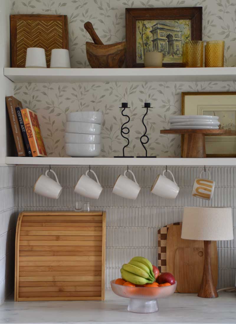 A pair of open kitchen shelves styled with plates, a cake stand, books, glassware, a wooden pestle and mortar and wavy candlesticks. Beneath is a faux marble worktop with a bread bin, a wood-based lamp and a glass bowl of fruit, and mugs are hanging from the lower shelf.