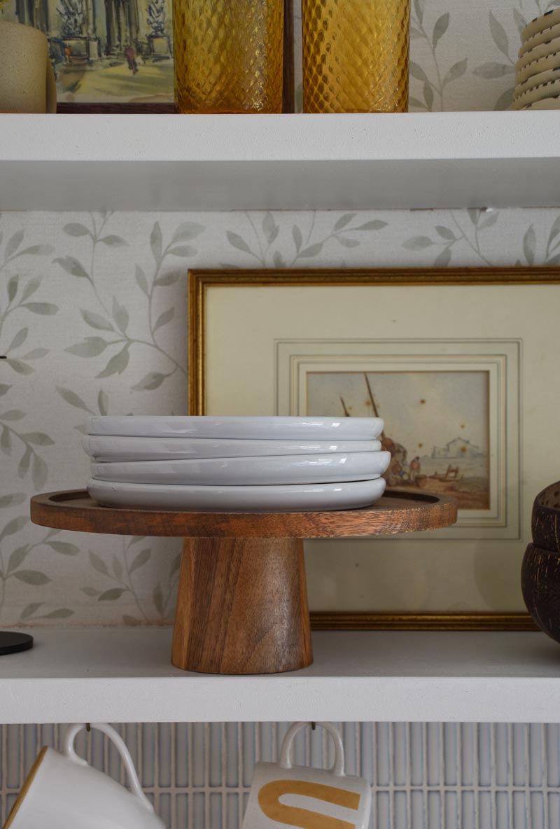 A stack of white plates sits on a wooden cake stand on an open kitchen shelf. The wall behind is papered in a soft leaf print, and mugs and glassware are just visible above and below.