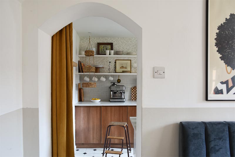 A peek into the kitchen through a mustard yellow thick ribbed curtain. Open shelving with plates, glassware, a cake stand and cutting boards sits above a faux marble worktop. A painting of a Black woman and the top of a banquette seat are just visible in the foreground.